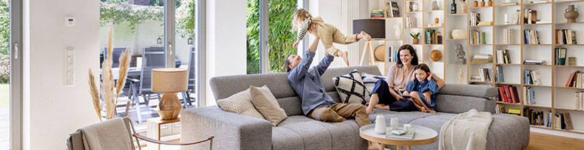 Gezin geniet in lichte woonkamer, vader tilt kind op, moeder en dochter op grijze bank in modern smart home