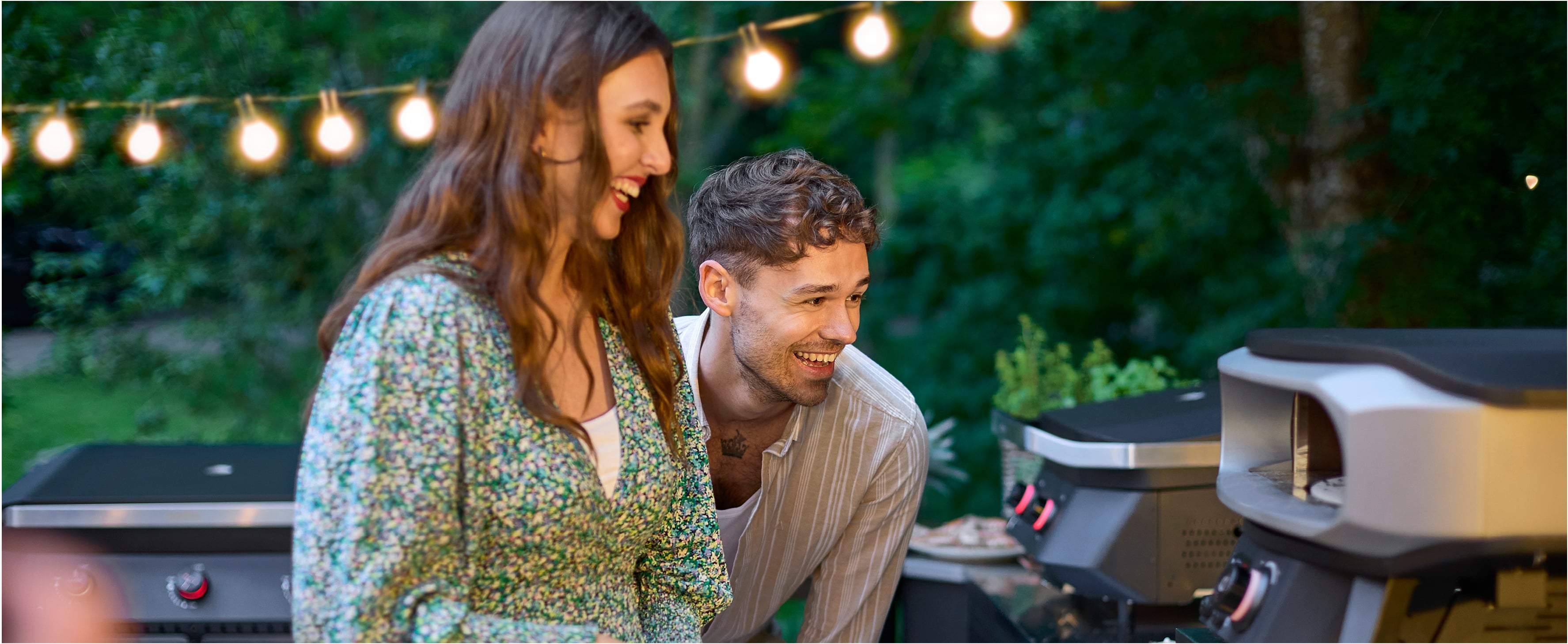 Lachend stel geniet van zomeravond bij Cozze pizzaoven en gasbarbecue in de tuin – gezellige sfeer met lichtjes