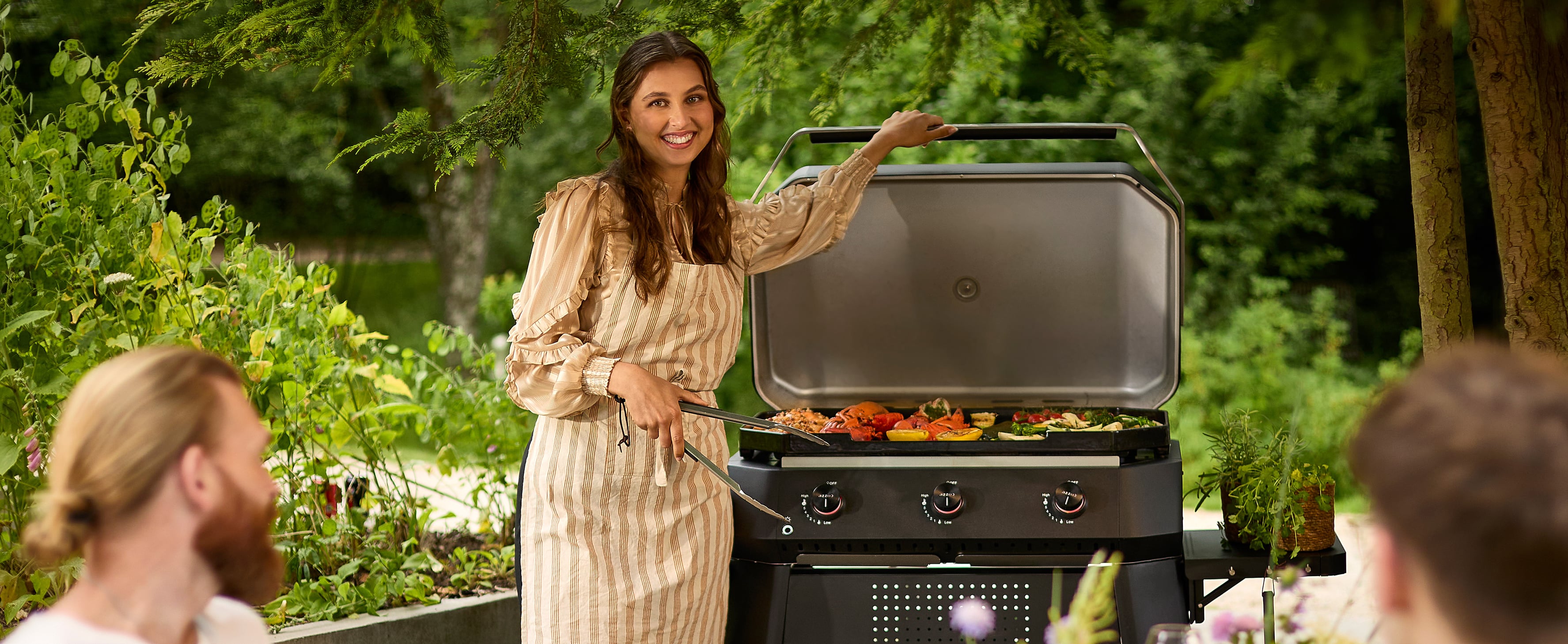 Lachende vrouw in schort grilt kleurrijke groenten op Cozze gasbarbecue met 3 branders in de tuin – zomerse sfeer met vrienden