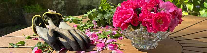 Tuinhandschoenen en een tuinschaar liggen buiten op een ronde houten tafel met een vaas met roze bloemen.