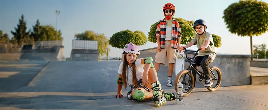 Drie kinderen met helmen en beschermingsuitrusting van Wozinsky op skateboard, rolschaatsen en BMX-fiets in skatepark